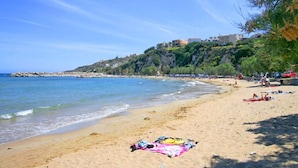 Ein Strand mit klarem, blauem Wasser, einem sandigen Ufer und Gebäuden an einem Hang im Hintergrund.