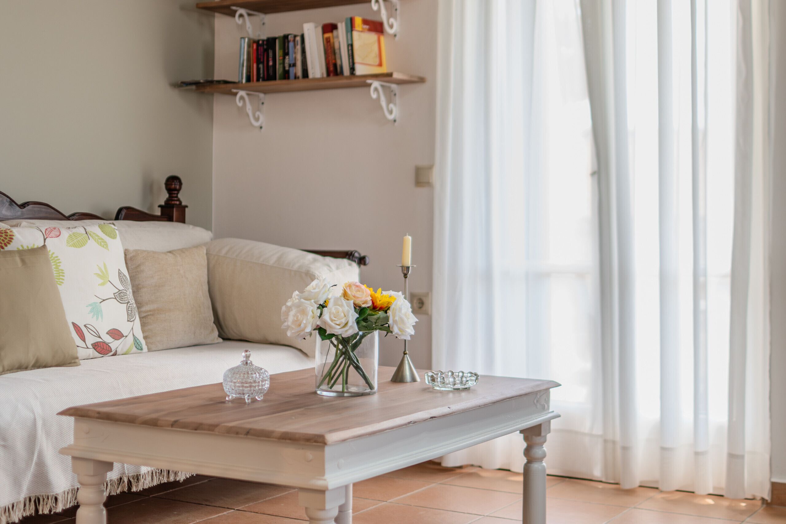 Un salón ordenado con un sofá, una mesa de centro, un jarrón con flores, una vela y una estantería.