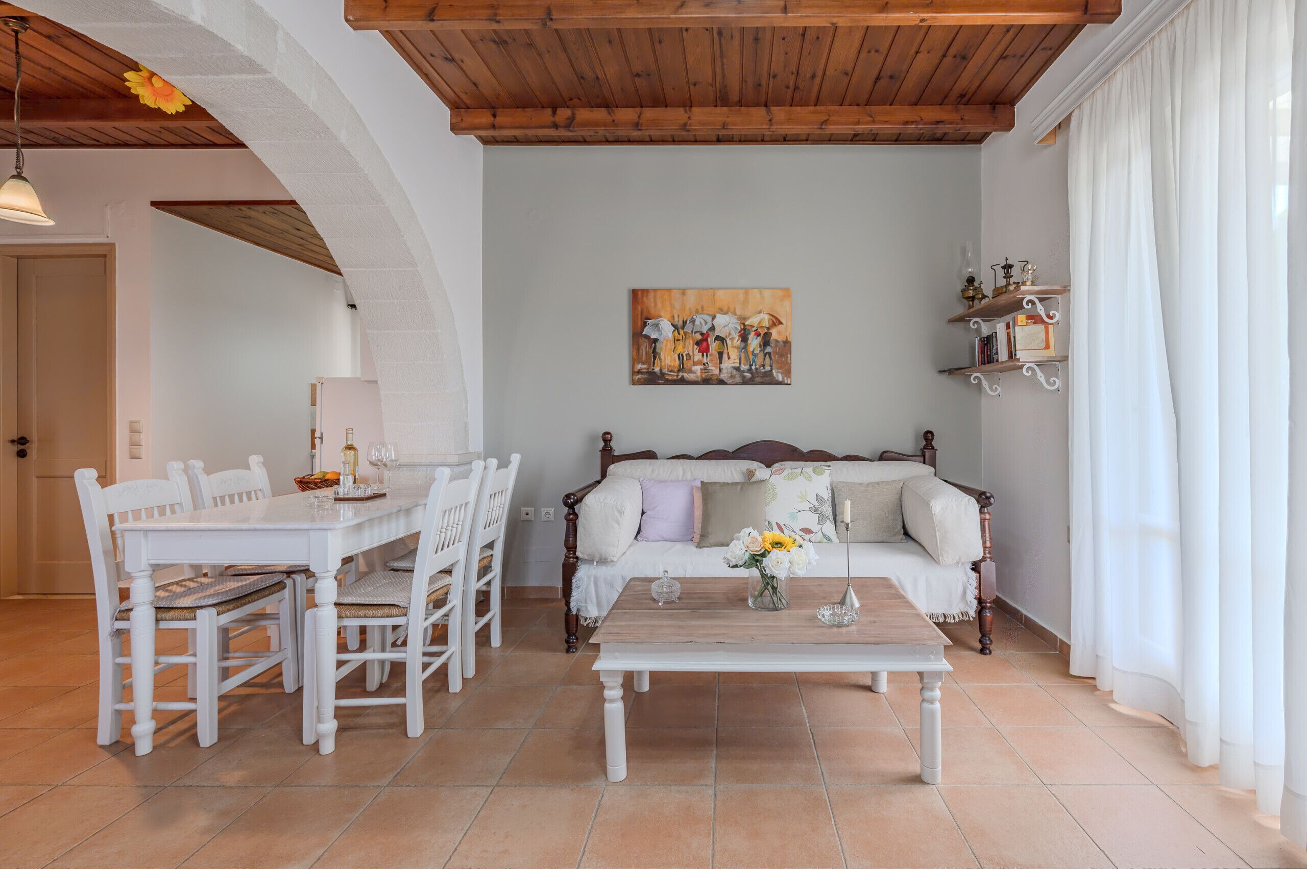 A dining area with a white table and chairs, a wooden coffee table with a vase of flowers, a sofa with cushions, and a painting on the wall.