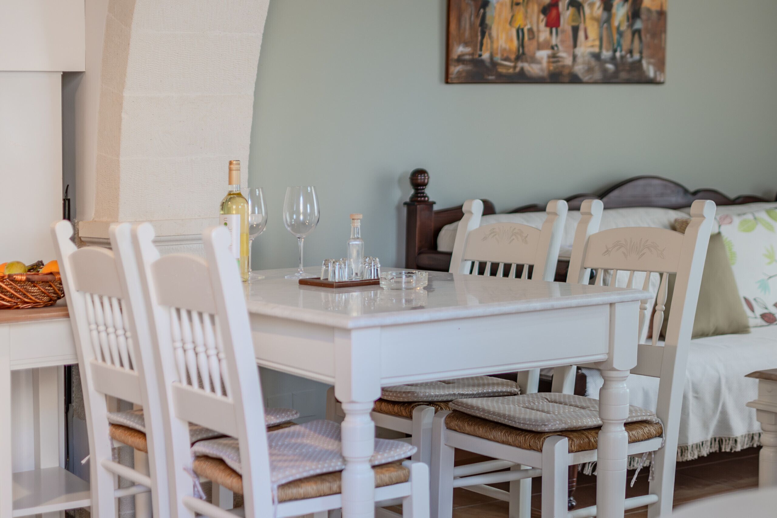 A dining area with a white table and chairs, a bottle of wine, and glasses. There is a bed with a headboard and a painting on the wall.