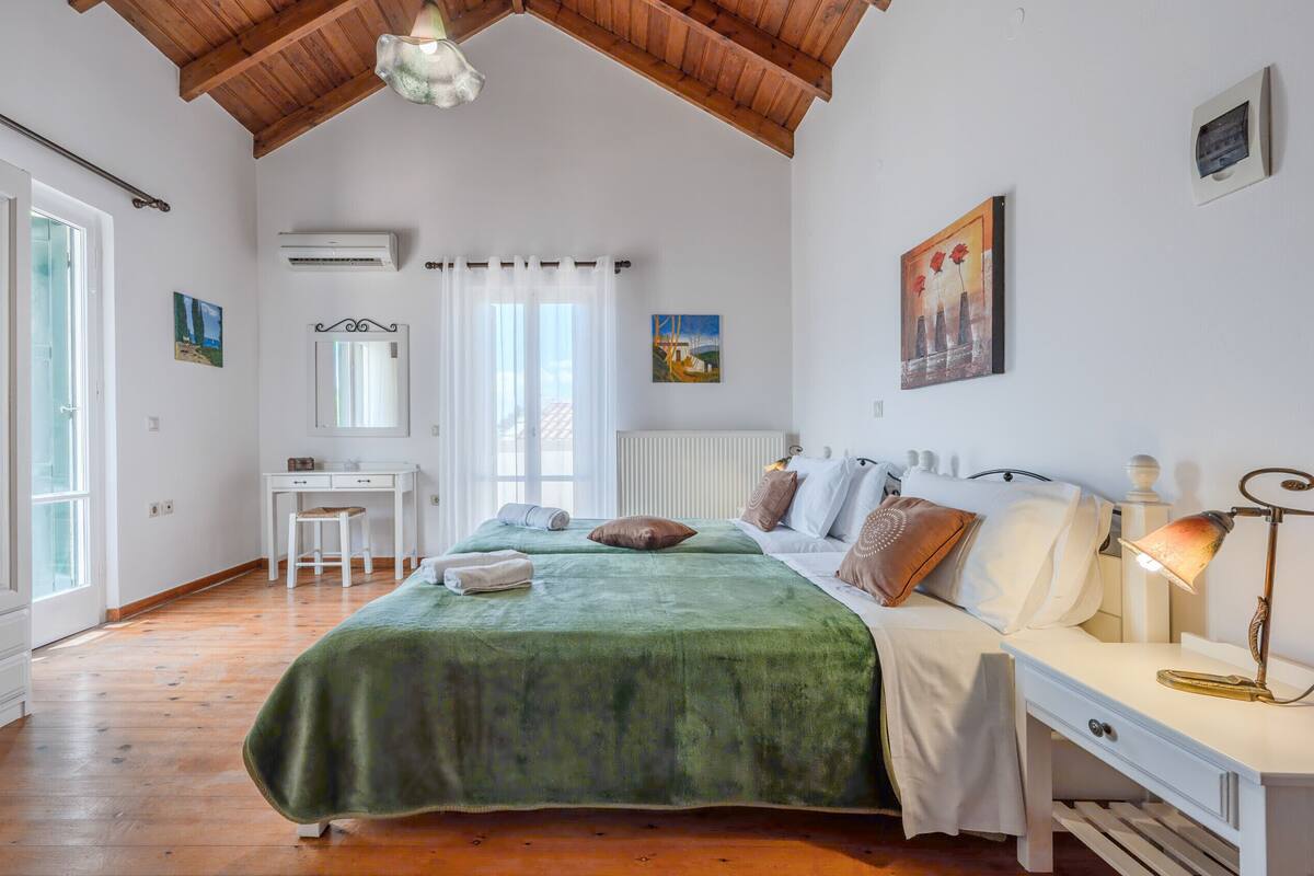 A bedroom with a wooden ceiling, a bed with a green bedspread, a desk with a chair, a radiator, and a window with curtains.