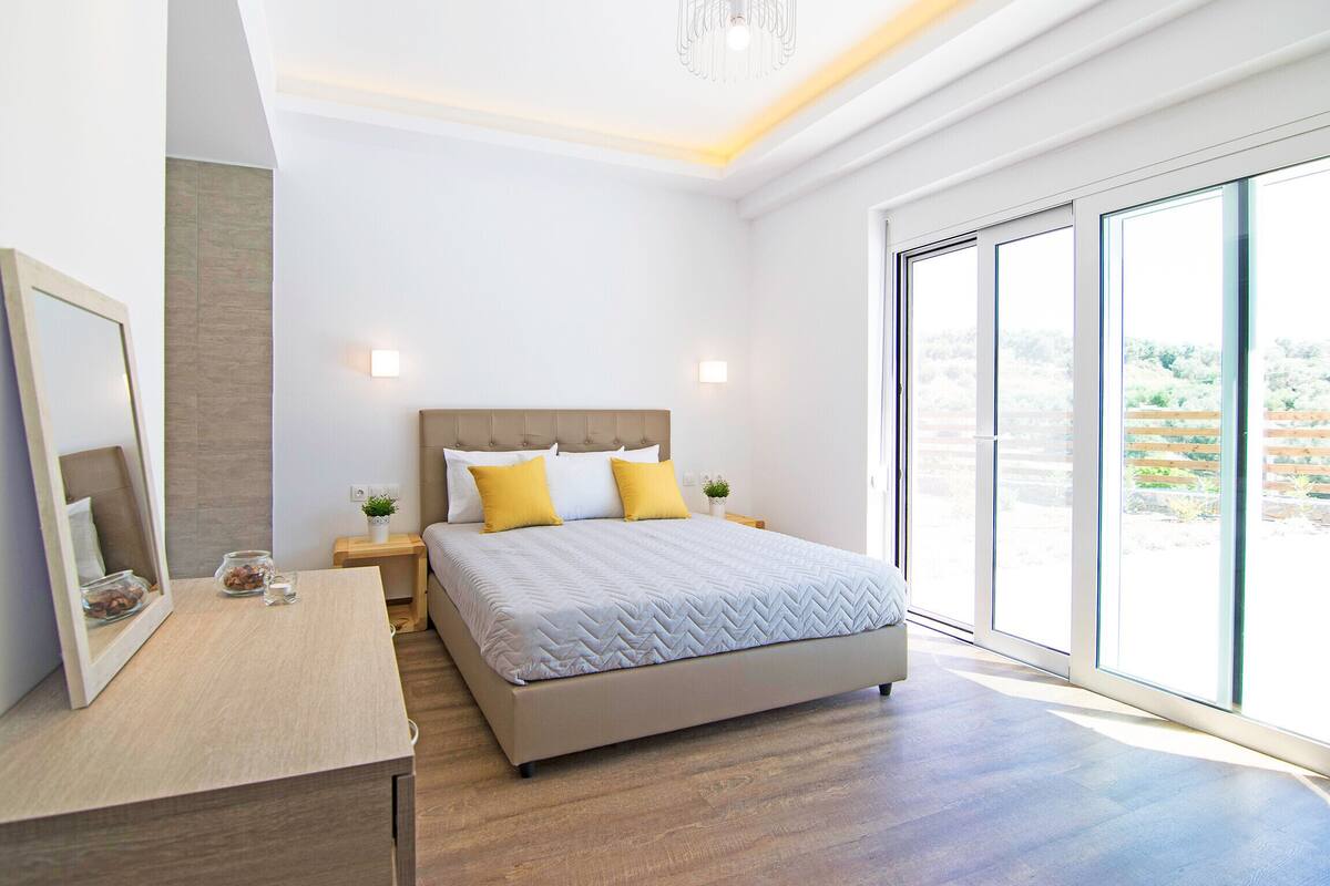A modern bedroom with a large bed, a bedside table, a mirror, and a view through large windows.