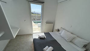 A hotel room with a bed, a nightstand, a window with a view of buildings, and a door leading to an outdoor area. - Villa Verdi Mykonos (mykonos)