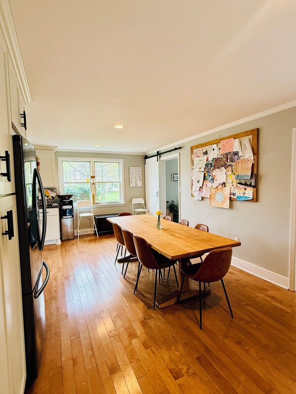Un amplio comedor con una mesa y sillas de madera, una cocina al fondo y un tablón de anuncios con varias notas e imágenes en la pared.