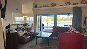 Un salón espacioso con un sofá rojo, una mesa de centro y un comedor con candelabro. Amplias ventanas ofrecen vistas del exterior.