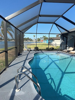 A covered swimming pool with a clear view of the surrounding area, including a grassy lawn and a body of water. - Canal-Front Home, Heated Pool,  75” TV , Bikes (Cape Coral)