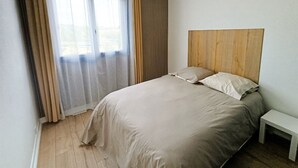 A bedroom with a bed, a bedside table, a wooden headboard, and a window with curtains.
