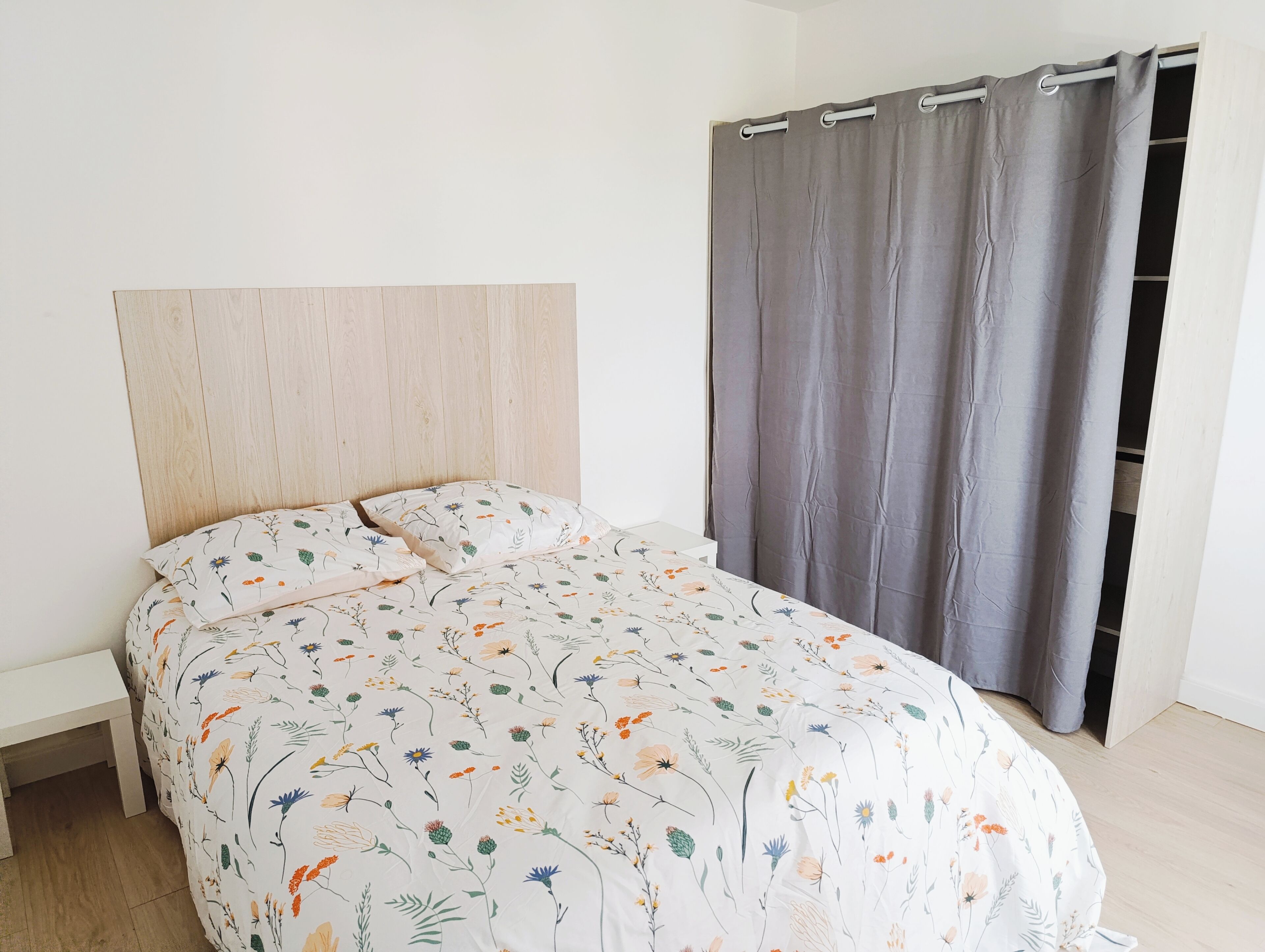 A bed with a floral bedspread, a wooden headboard, and a grey curtain.