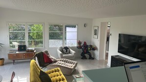 A living room with a sofa, armchair, and a TV. There are windows with a view of greenery.