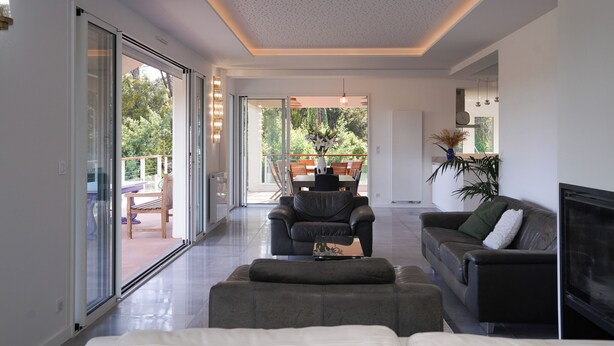A modern living room with a sofa, a coffee table, and a TV. There is a sliding glass door leading to an outdoor patio area with a dining table and chairs. - The Beach House (Seignosse)