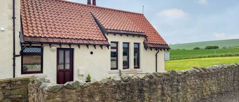 A house with a red-tiled roof, stone wall, and a grassy field in the background.