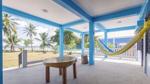 Un porche techado con una mesa y sillas, una hamaca y vista a la playa y palmeras.