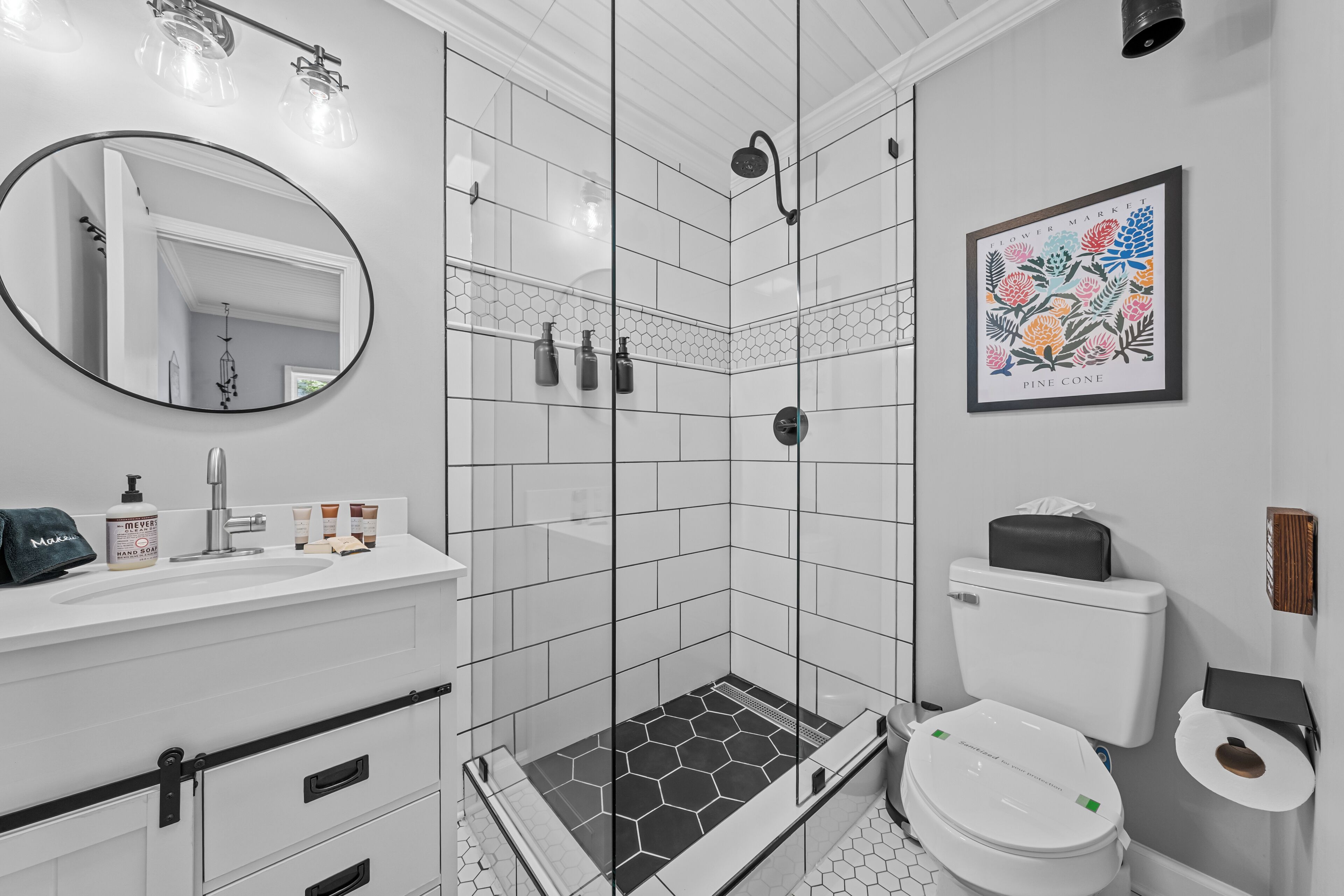 Een moderne badkamer met een douchecabine van glas, witte tegels, een ronde spiegel, een wastafel met toiletartikelen en een toilet met toiletrolhouder.
