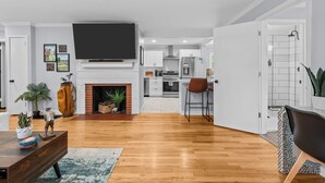 A modern living room with a fireplace, wooden flooring, a flat-screen TV, and a kitchen in the background. - Charming Downtown Corner Cottage - 1 Block Walk to Food & Fun - Dog Friendly! (Southern Pines)