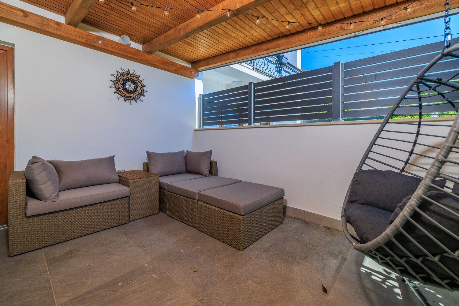 Ein überdachter Patio-Bereich mit einem Rattansofa, einem Hängesessel und einer dekorativen Wanduhr.