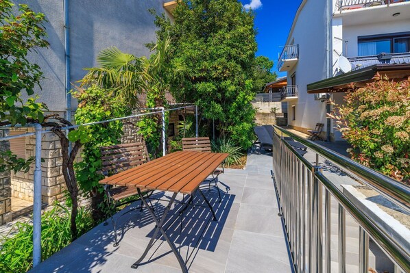 A sunny courtyard with a wooden table and chairs, surrounded by lush greenery and a stone wall.