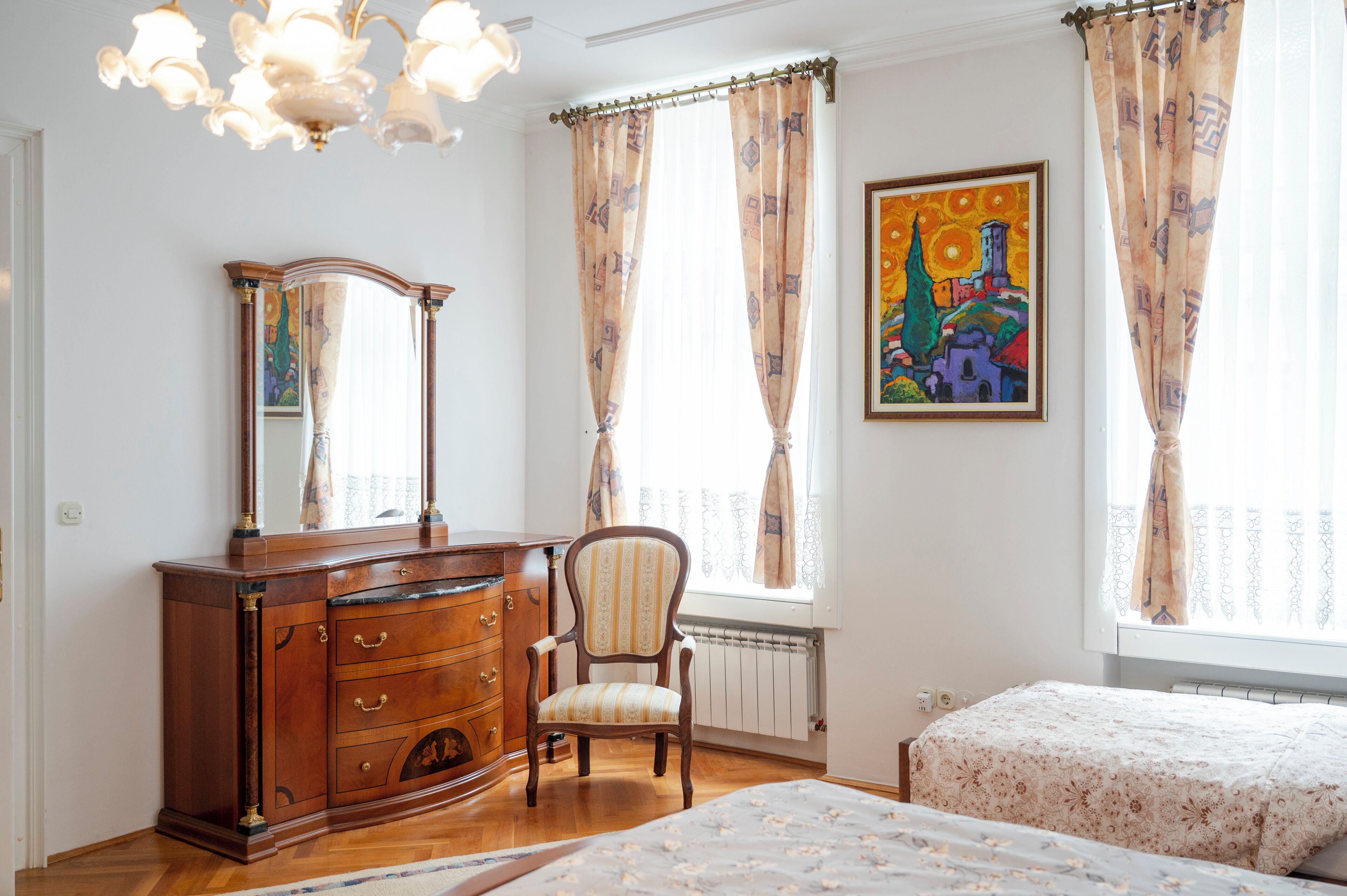 Un dormitorio con un aparador de madera, una silla, una cama con colcha estampada, un radiador y un cuadro en la pared.