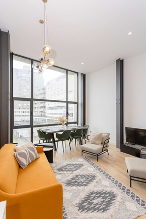 Un salon moderne comprenant un canapé, une table à manger avec des chaises, un téléviseur et de grandes fenêtres donnant sur la ville.