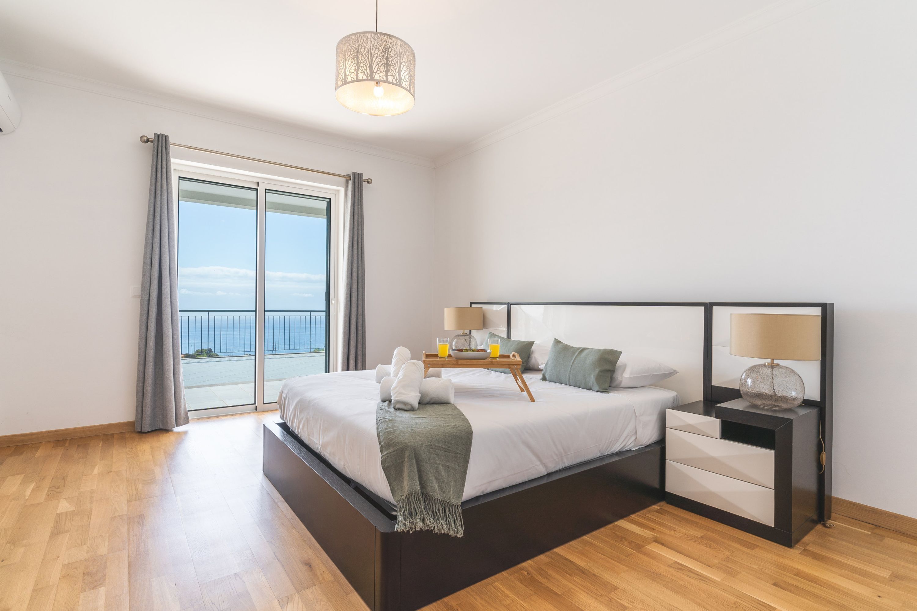 A modern bedroom with a large bed, bedside tables, and a view of the sea through a sliding door.