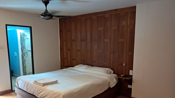 Egy hálószoba, melyben fából készült ágyvég található, az ágyon fehér ágynemű van, emellett van egy éjjeliszemély és egy mennyezeti ventilátor.