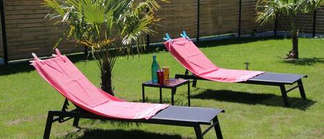 Deux chaises longues avec des serviettes rouges, une petite table avec des boissons et un palmier dans un espace extérieur verdoyant.