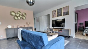 A modern living room with a blue sofa, a flat-screen TV, decorative wall art, and a small table with glasses and a bottle.
