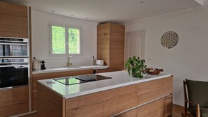 A modern kitchen with a central island, wooden cabinets, a built-in oven, and a window providing natural light. - Air-conditioned villa with swimming pool between the sea and the Cevennes mountains. (La Calmette)