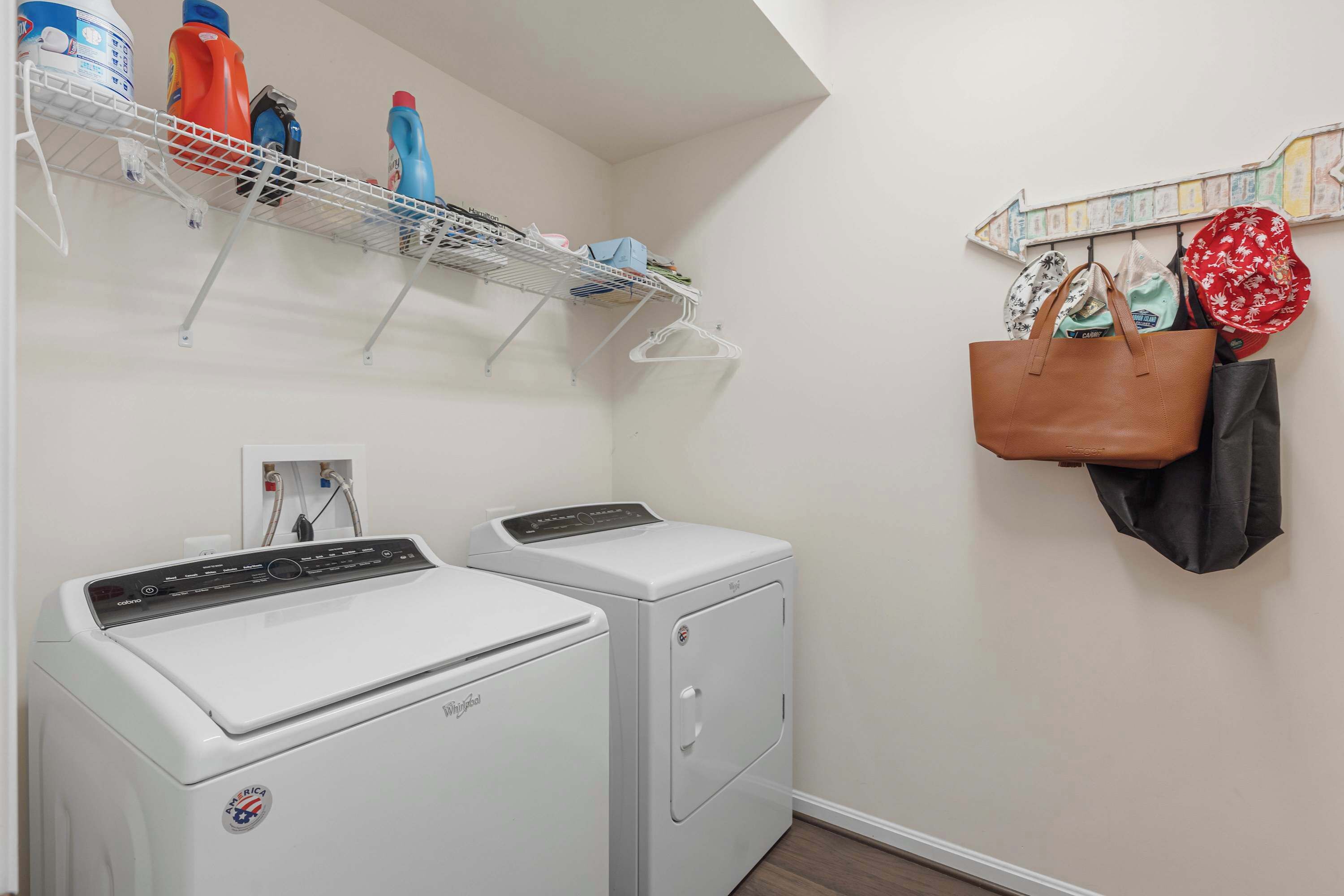 A laundry area with two white front-loading washing machines, a metal shelf above holding bottles and hangers, and a wall-mounted rack with bags and hats.