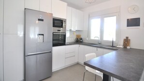 A modern kitchen with white cabinets, stainless steel appliances, and a dining area with a dark countertop. - Zennova 29 Vourvourou Garden House (Vourvourou)