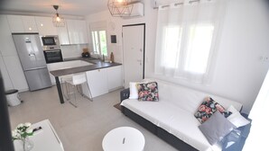 A modern kitchen with stainless steel appliances, a dining area with a white table and chairs, and a living area with a sofa and cushions. - Zennova 29 Vourvourou Garden House (Vourvourou)