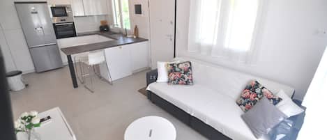 A modern kitchen with stainless steel appliances, a dining area with a white table and chairs, and a living area with a sofa and cushions.