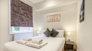 A neatly made bed with folded towels, a bedside table with a lamp, and a window with a patterned blind.