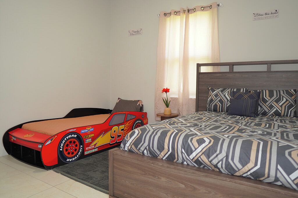 Ein Schlafzimmer mit einem Bett, einem Nachttisch, einem roten Spielzeugauto und einem Fenster mit Vorhängen.