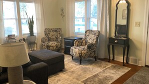 Un salon avec deux fauteuils à motifs, un pouf foncé, une lampe sur pied, une petite table d’appoint et un miroir posé sur une commode.