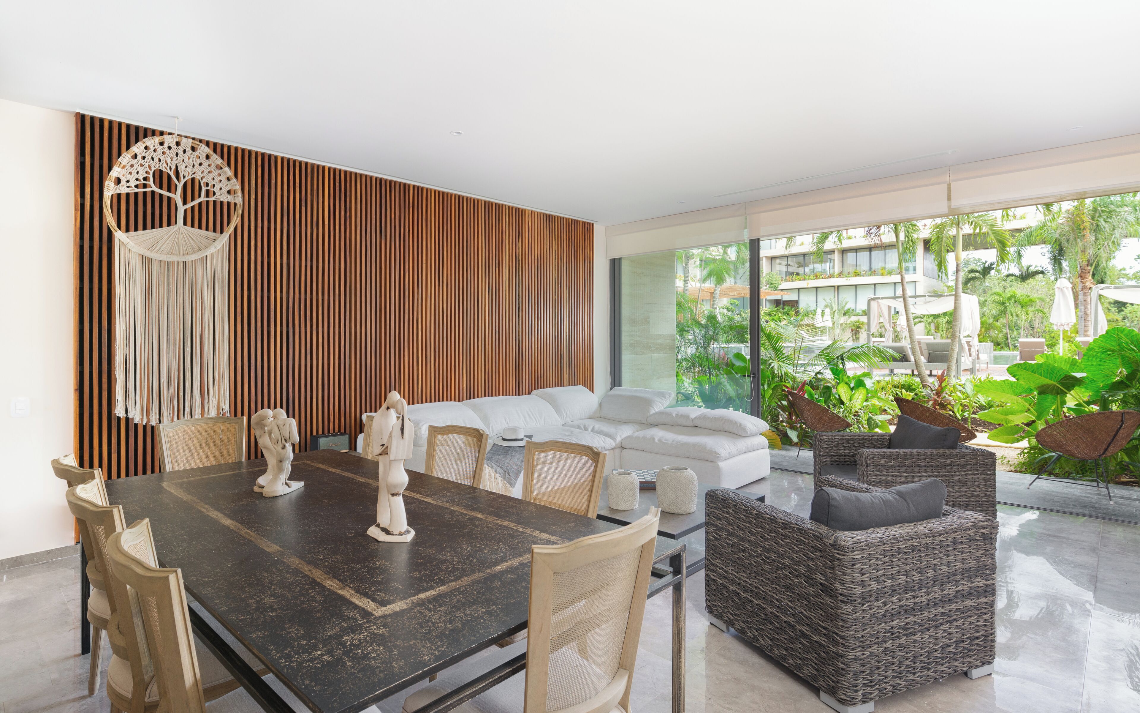 A modern living room with a dining area, featuring a dark wooden table, wicker furniture, and a large window overlooking a lush garden.