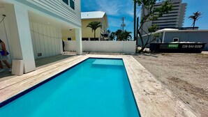 En swimmingpool med betonfliser, omgivet af en hvid mur og en bygning med gul facade.
