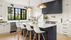 A modern kitchen with a large island, white cabinetry, and a black accent wall. There are three bar stools around the island and a large window providing natural light. - Ocean Whisper Retreat (Victoria)