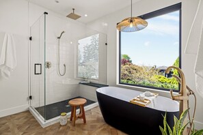 Ein modernes Badezimmer mit einer begehbaren Glasdusche, einer freistehenden Badewanne und einem großen Fenster mit Blick auf Grünflächen.