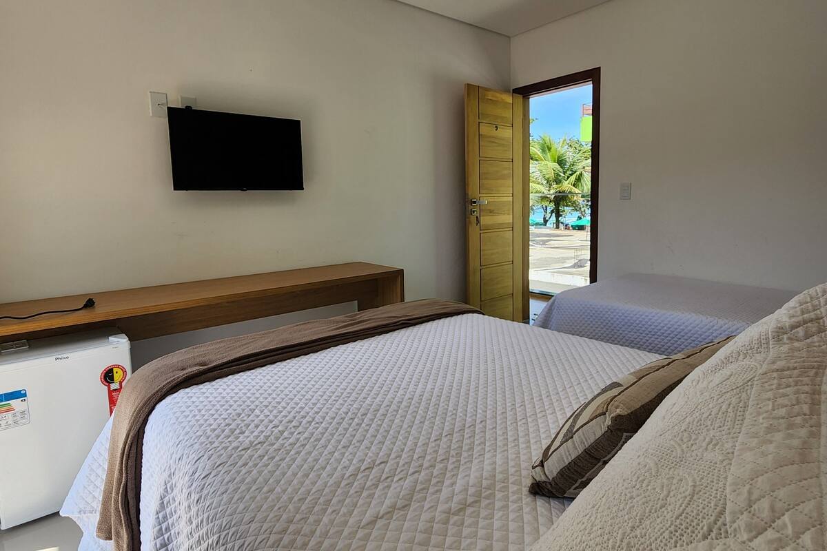 A bedroom with a bed, a TV mounted on the wall, a wooden bedside table, and a window with a view of palm trees.