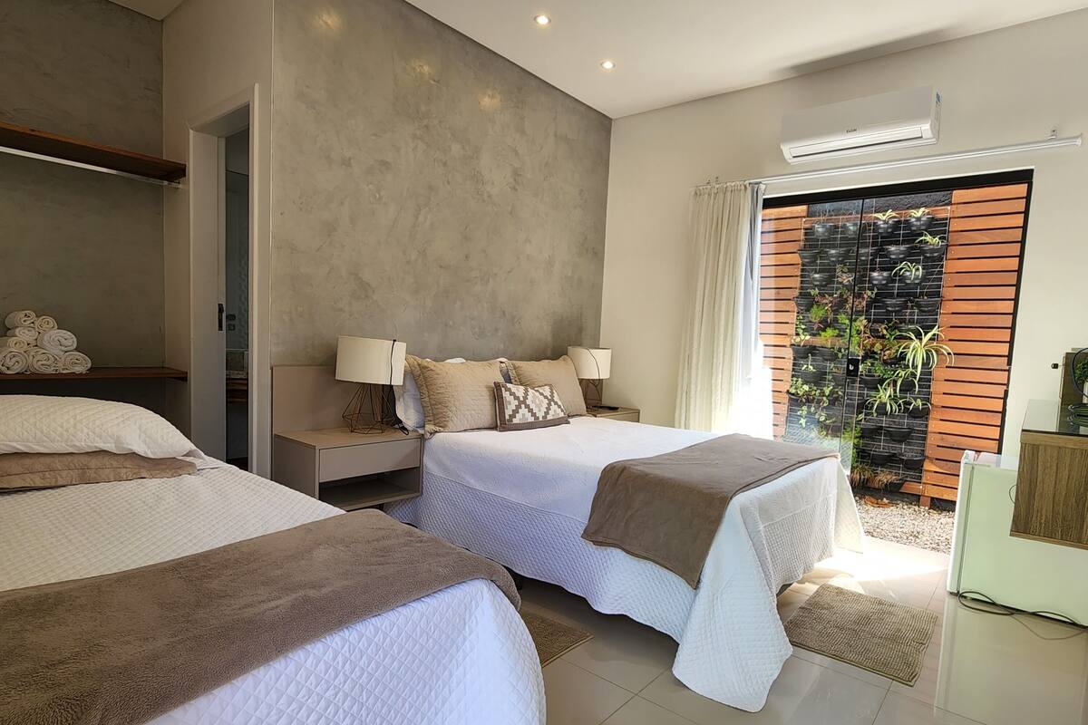 A modern hotel room with two beds, a large window with a view of a garden, and a ceiling with recessed lighting.