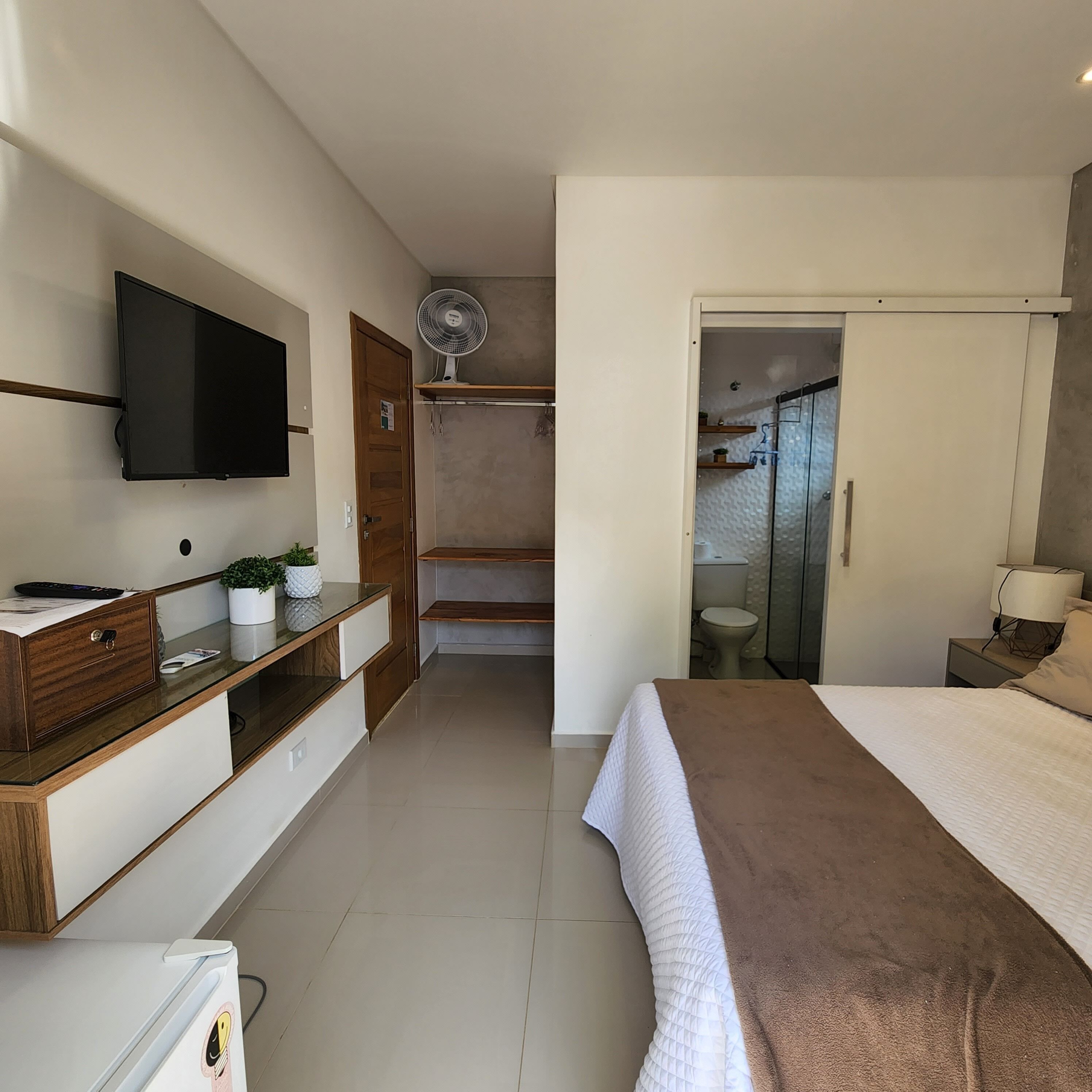 A bedroom with a bed, a TV mounted on the wall, a desk with a computer, a fan, and a bathroom visible through an open door.