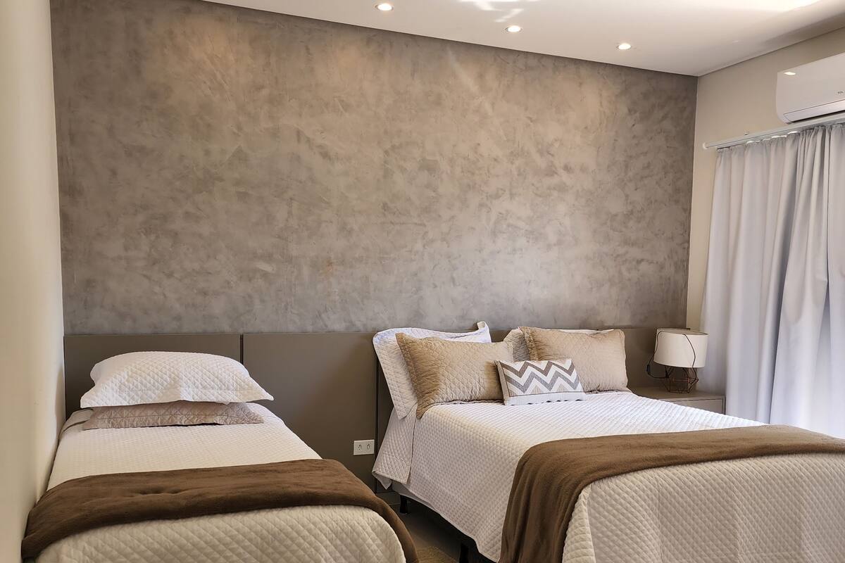 A hotel room with two beds, a large window with curtains, a bedside table with a lamp, and a wall-mounted air conditioning unit.