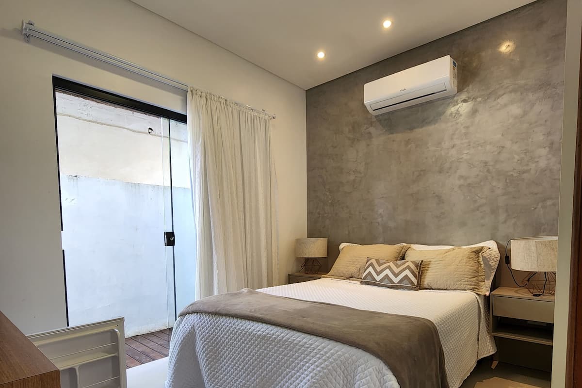 A modern bedroom with a bed, bedside lamps, a built-in air conditioner, and a sliding glass door.