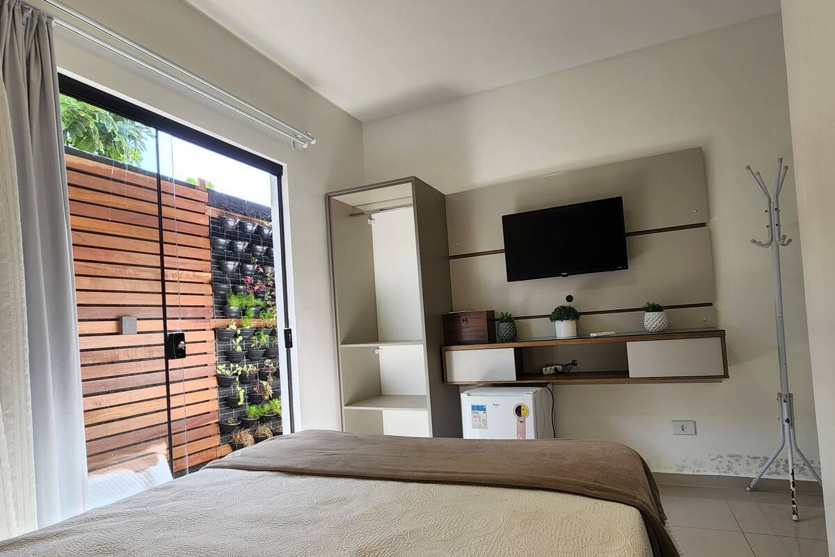 A modern bedroom with a large bed, a TV mounted on the wall, and a sliding glass door leading to a balcony with plants.
