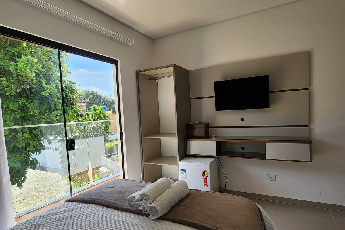 A modern bedroom with a large bed, a flat-screen TV mounted on the wall, and a sliding glass door leading to a balcony with a view of greenery.