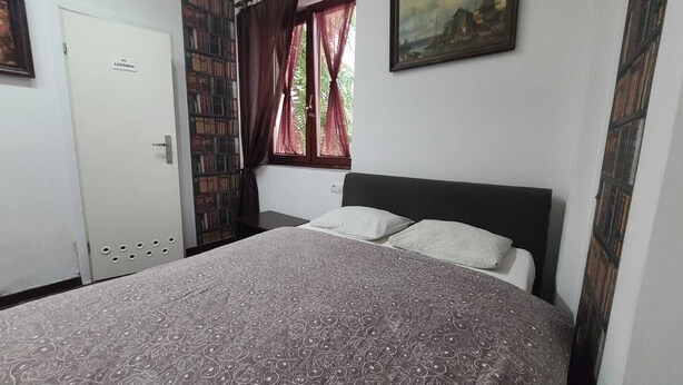 Camera da letto con un letto, due cuscini, una libreria, una porta con la scritta 'WC' e una finestra con tende.