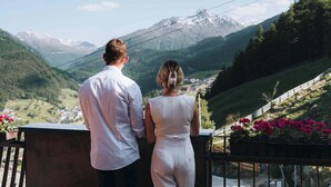 A couple standing on a balcony overlooking a mountainous landscape with a village below. - Grünwald Resort Sölden (Soelden)