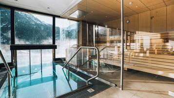 Una piscina interna moderna con barriera in vetro, rivestimenti in legno e vista sulle montagne innevate.
