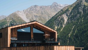 Un chalet moderne en bois, doté de grandes fenêtres et d’un balcon, qui se dresse sur un fond de montagnes.