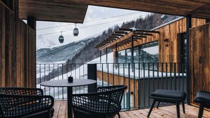 A wooden deck with a round table and chairs, a staircase leading to a glass balcony, and a view of snow-covered mountains.